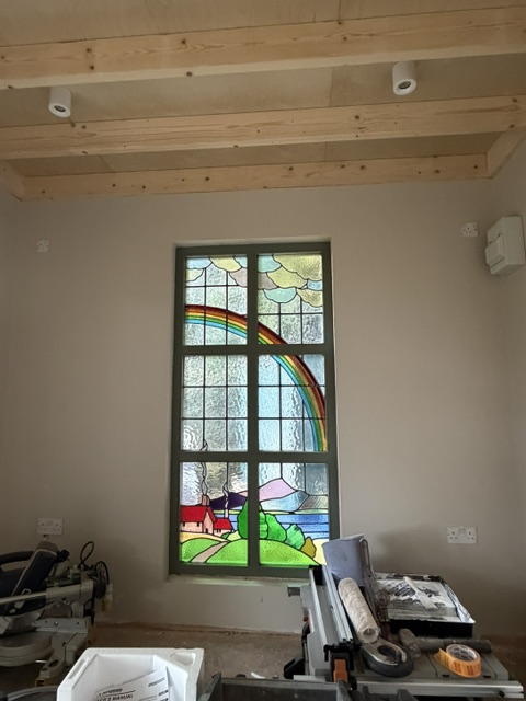 Repaired leaded stained glass window with a rainbow arching over a cottage and hills, seen from inside a timber-framed room
