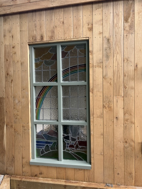 Repaired stained glass rainbow window in a sage green frame set into timber cladding, seen from the garden