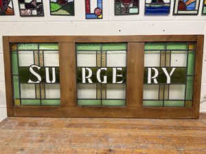 Restored stained glass panel in a wooden frame with the word SURGERY in white lettering on green and clear leaded glass